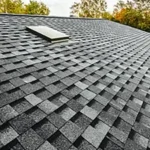 Black asphalt roof shingles installed on a sloped rooftop with a skylight, surrounded by trees - Asia Jaya Sepadu.